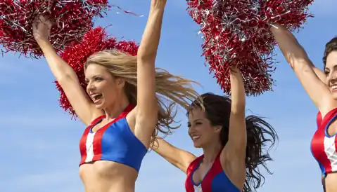 Cheerleaders performing with red and silver pom-poms under a bright sky.
